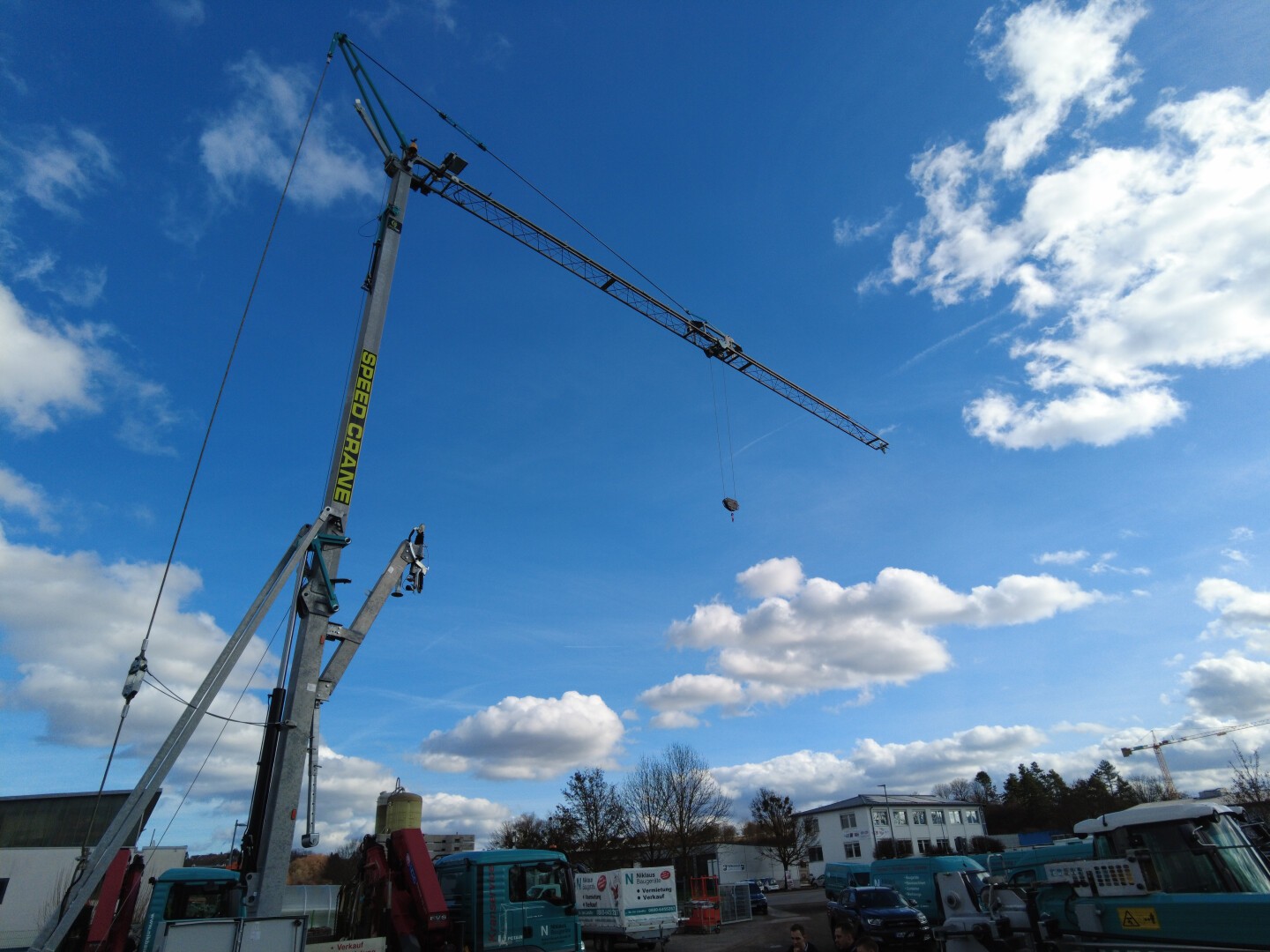 Vor einem strahlend blauen Himmel mit vereinzelten Wolken thront ein hoher Baukran über einer Baustelle mit Lastwagen und Ausrüstung.