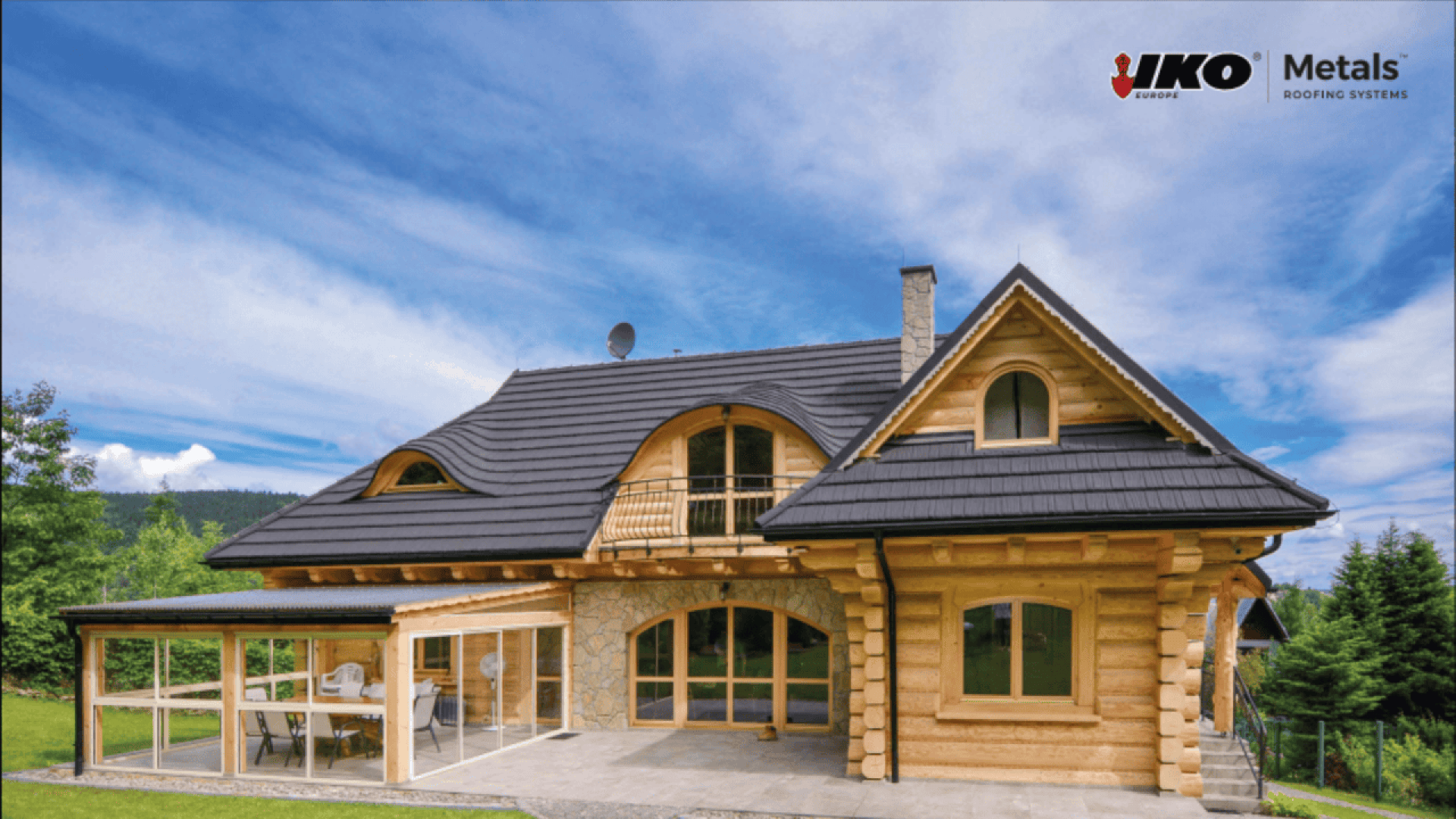 A wooden house with a modern black metal roof, large windows, and a covered patio area sits in a green landscape under a blue sky. The IKO Metals Roofing Systems logo is in the top right corner.