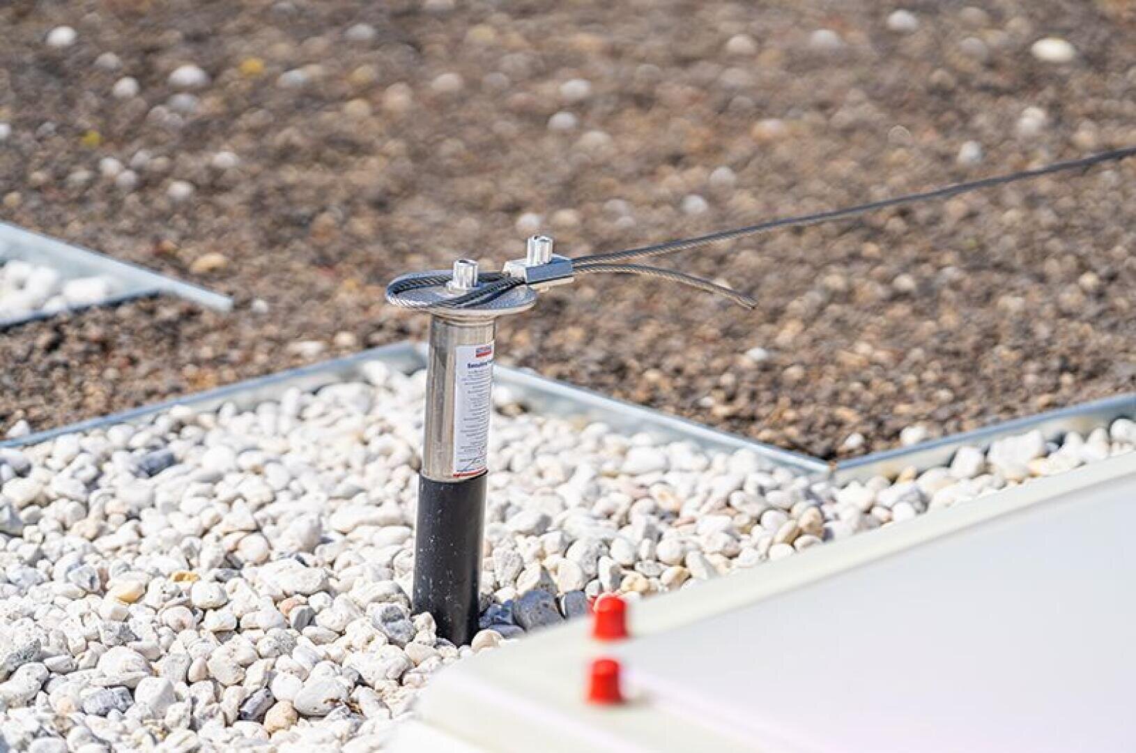 A metal anchor post with a cable attached to it is installed amidst white gravel and earth, and a few red fasteners and metal edges can be seen nearby.