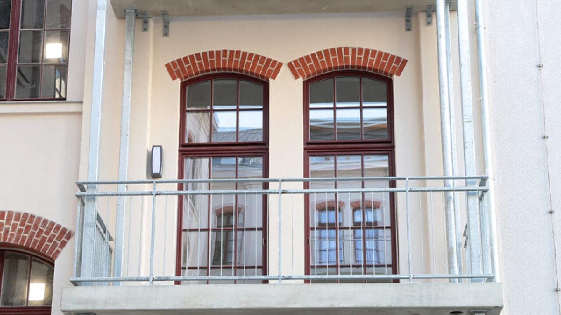 A small balcony with a metal railing in front of two tall arched windows framed with red brick on a light-colored building. The windows reflect another building.