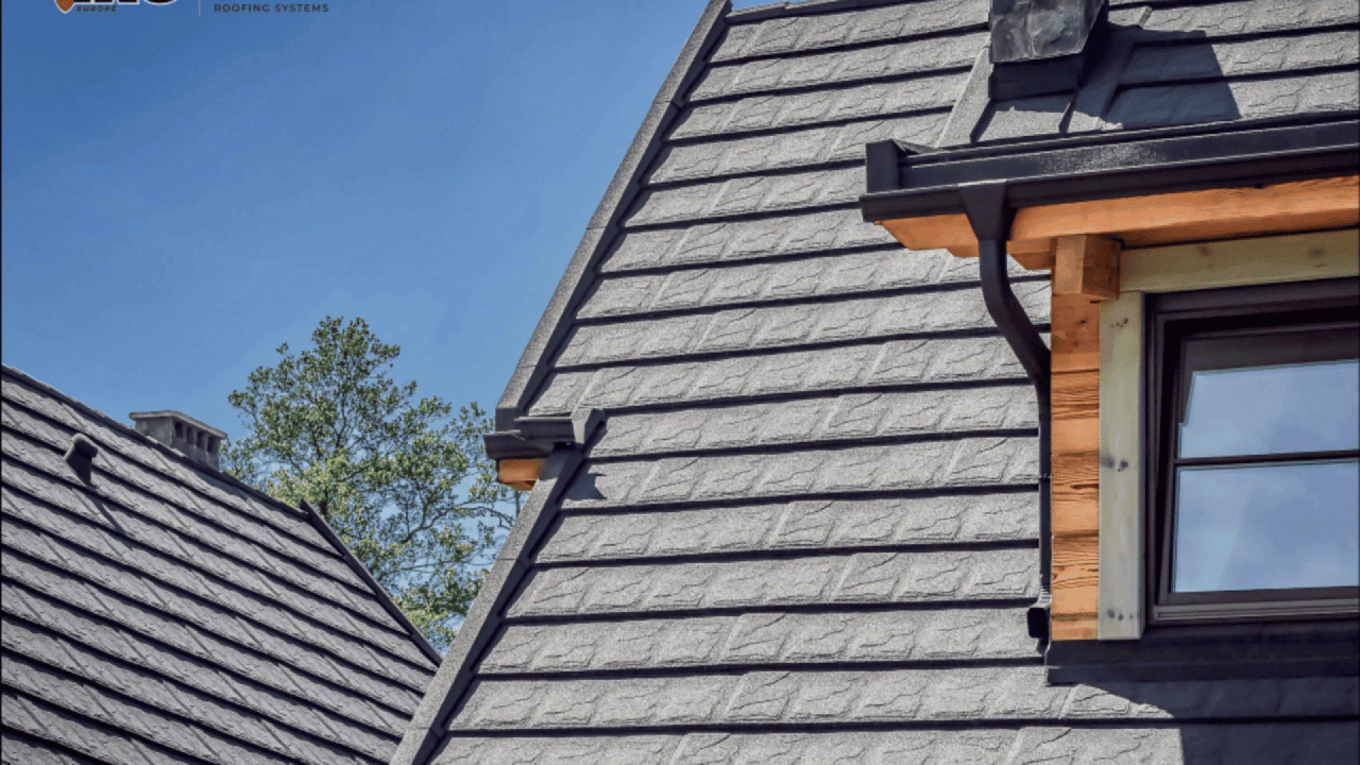 Close-up of a modern house roof with gray, textured shingles, black gutters, and a partially visible window with wood trim, under a clear blue sky. Trees can be seen in the background.