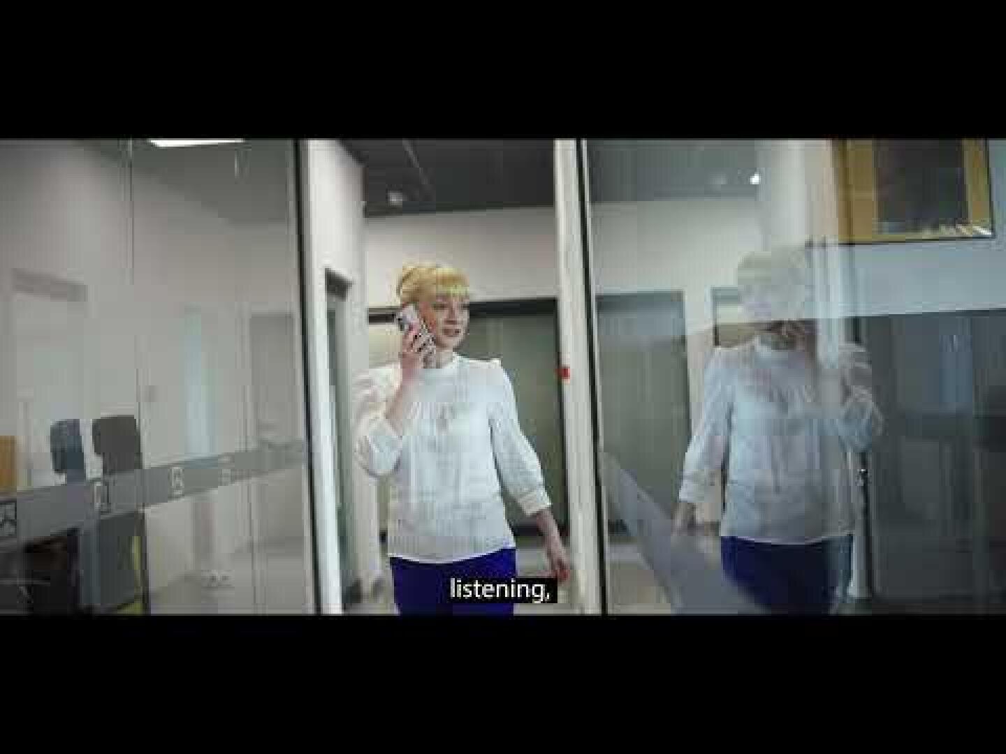 A person with short blond hair talks on a cellphone while walking past glass office walls; the caption on screen reads listening.