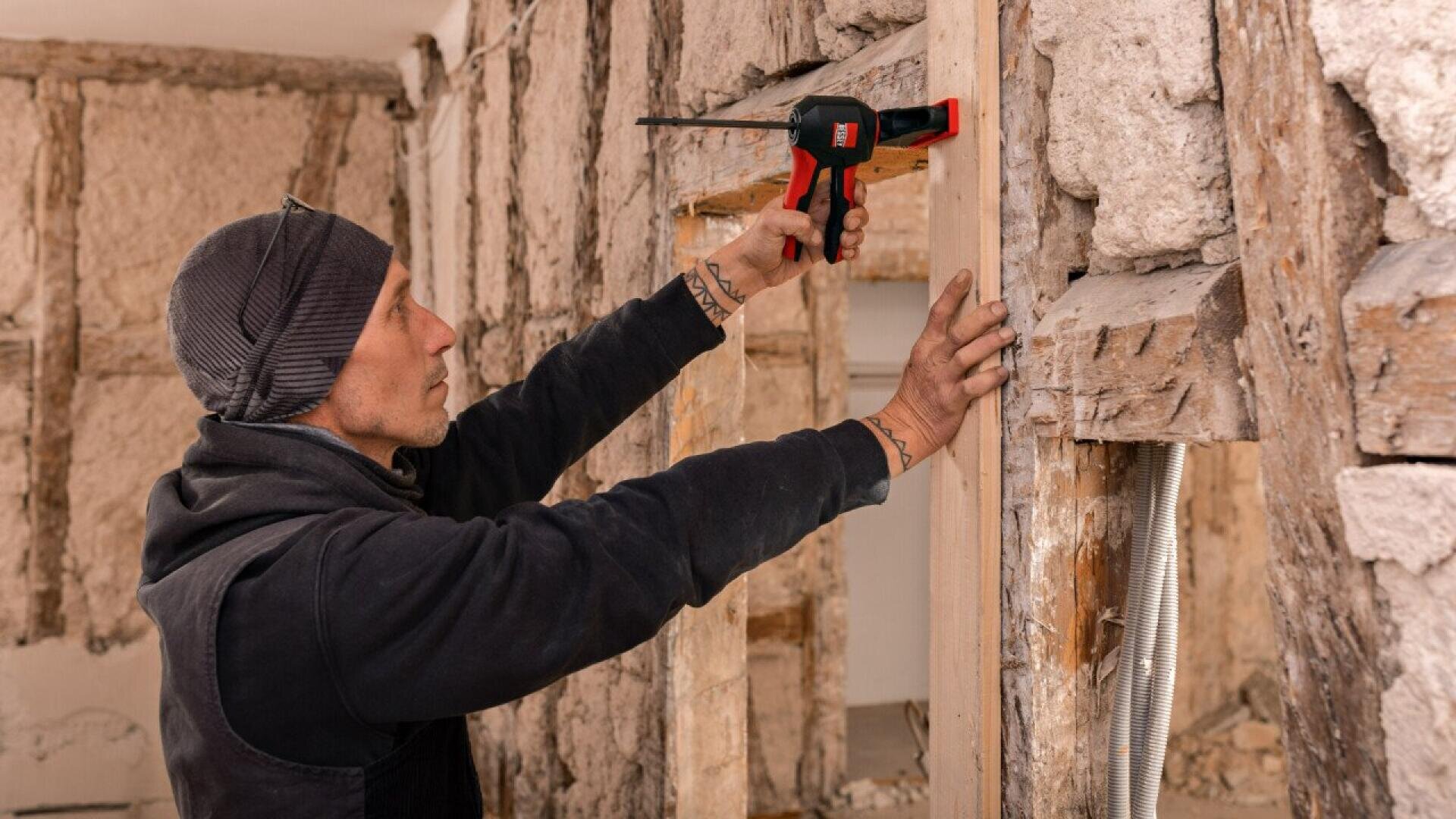 A person wearing a beanie and vest uses a power tool to secure a wooden beam inside a partially renovated room with exposed wooden studs and insulation.
