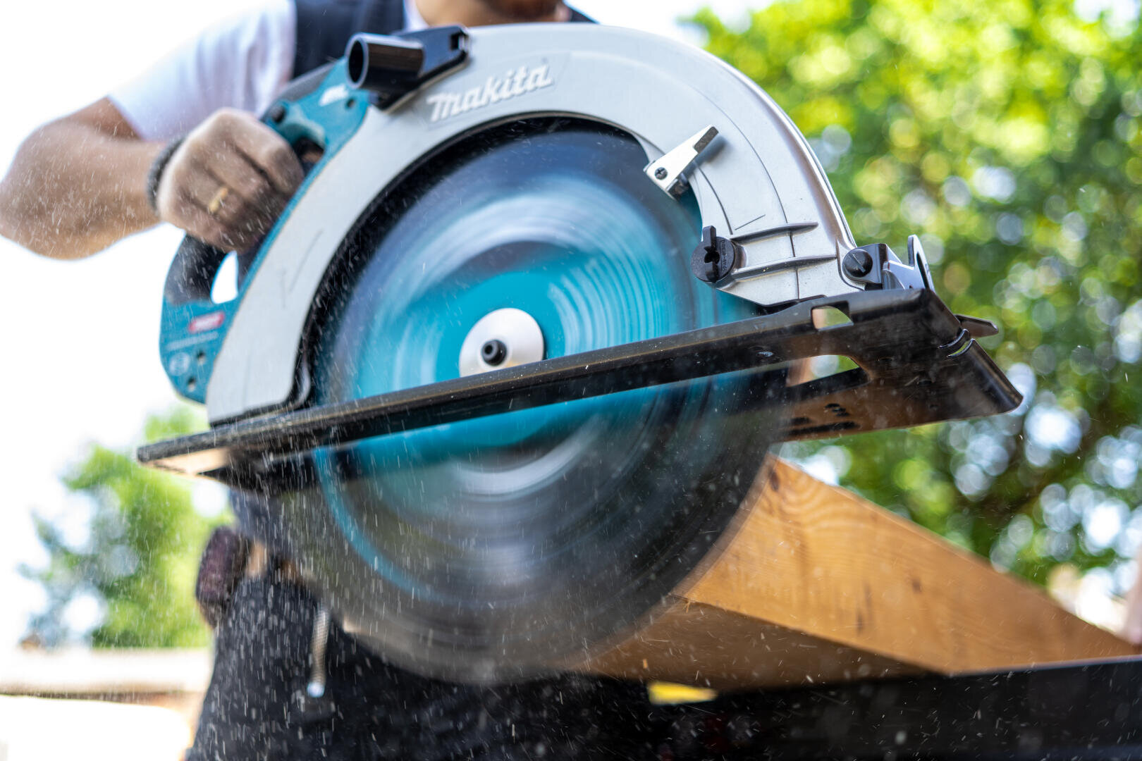 Eine Person schneidet mit einer Makita-Kreissäge ein Holzbrett im Freien. Das sich drehende Sägeblatt wirbelt Sägespäne auf, und im Hintergrund sind unscharfe grüne Bäume zu sehen.