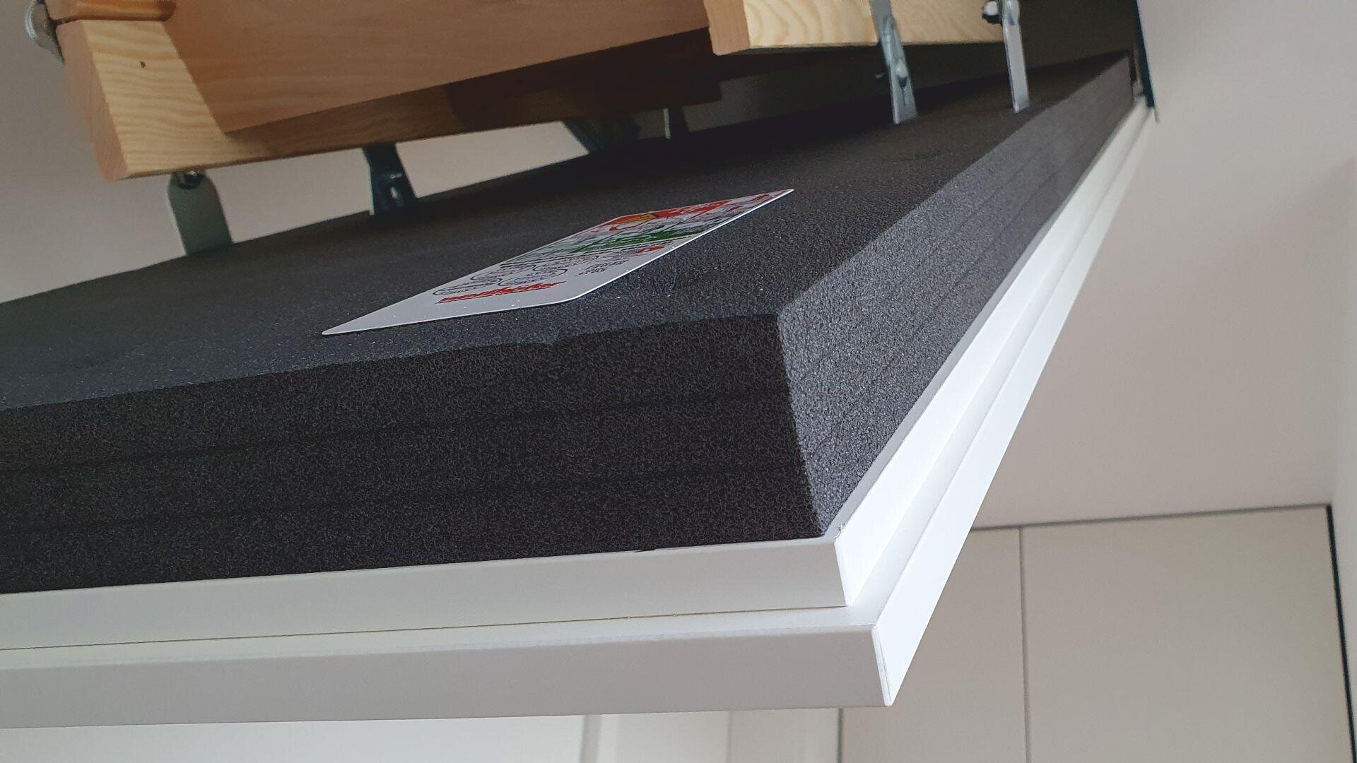 A close-up view from below of an insulated attic hatch door, partially opened, showing thick black foam insulation, a product label, and wooden attic stairs above.