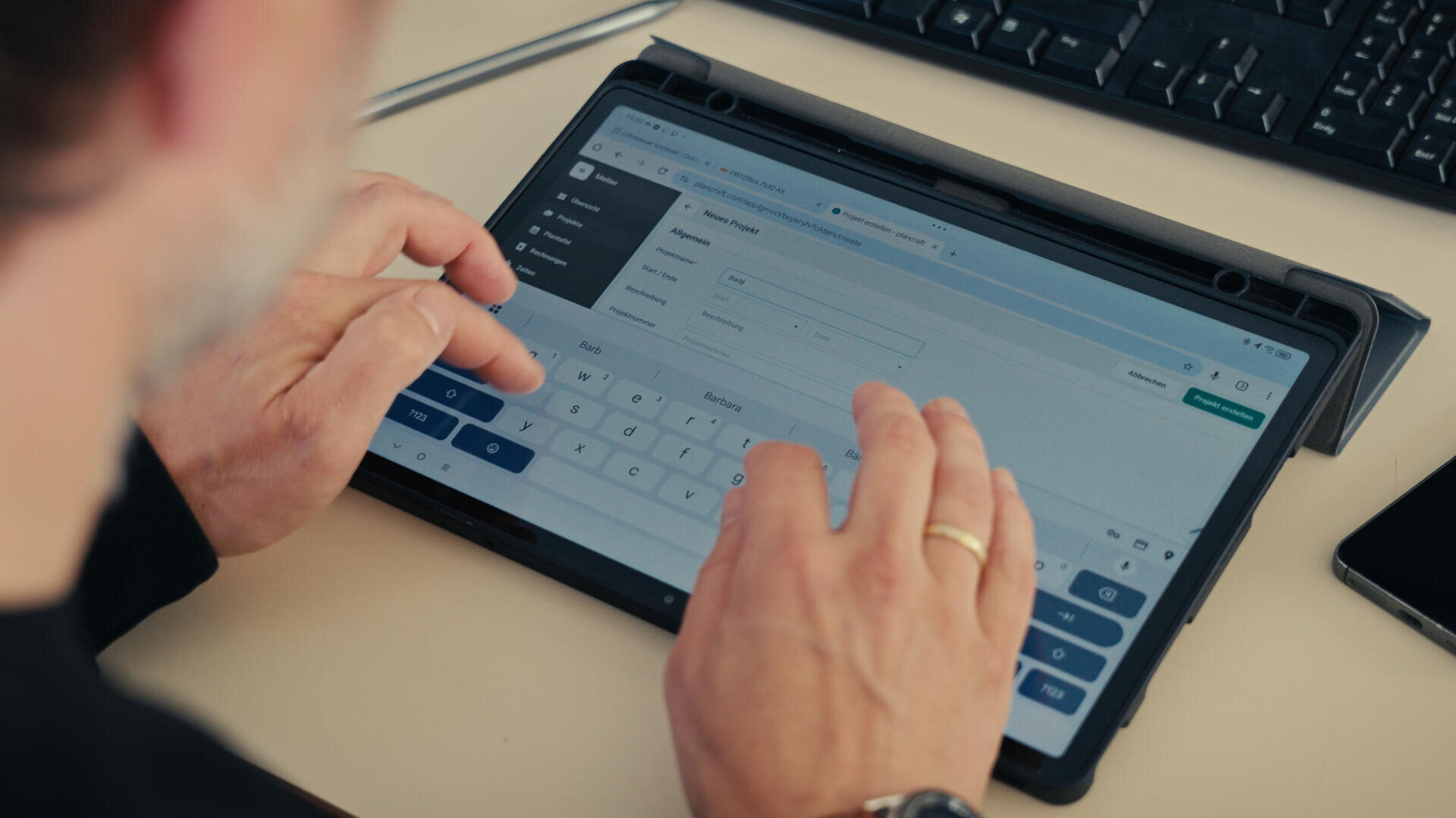 Eine Person tippt auf der Bildschirmtastatur eines Tablets und füllt ein Formular in einem Webbrowser aus. Das Tablet liegt auf einem Schreibtisch neben einer Tastatur und einem Stift. Die Hände der Person sind sichtbar, eine trägt einen goldenen Ring.