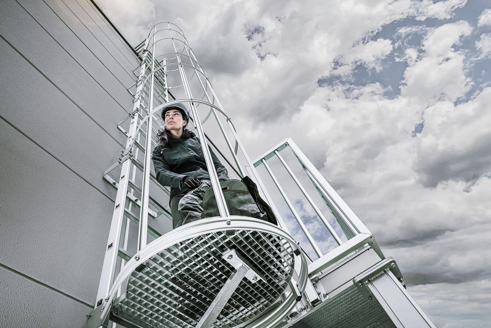 Eine Person mit Schutzausrüstung und Helm klettert eine Metallleiter hinauf, die an der Seite eines Gebäudes befestigt ist. Im Hintergrund ist ein bewölkter Himmel zu sehen.