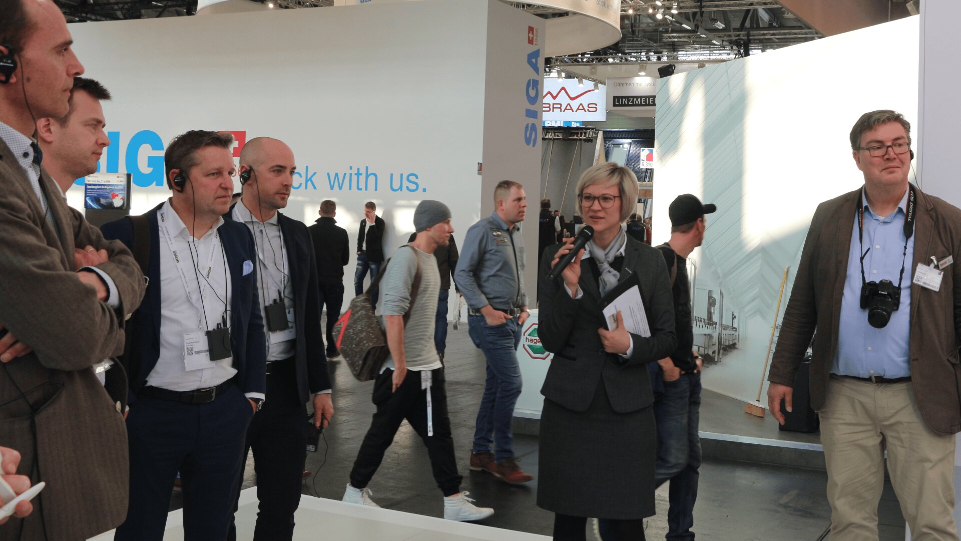 Eine Gruppe von Personen in Geschäftskleidung steht in einer Ausstellungshalle, eine Frau hält ein Mikrofon und spricht, während andere zuhören. Im Hintergrund sind mehrere Umstehende und ein großer weißer Stand zu sehen.