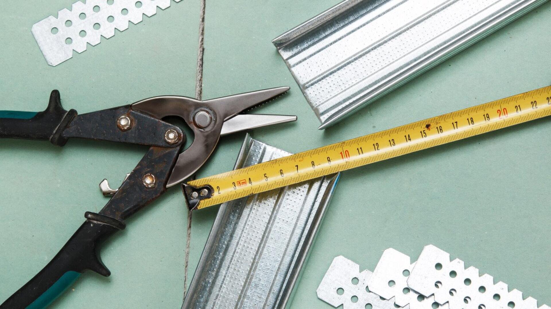 A pair of metal snips, a metal ruler measuring tape, metal corner beads, and metal plates are arranged on a green surface, suggesting tools and materials for construction or drywall work.