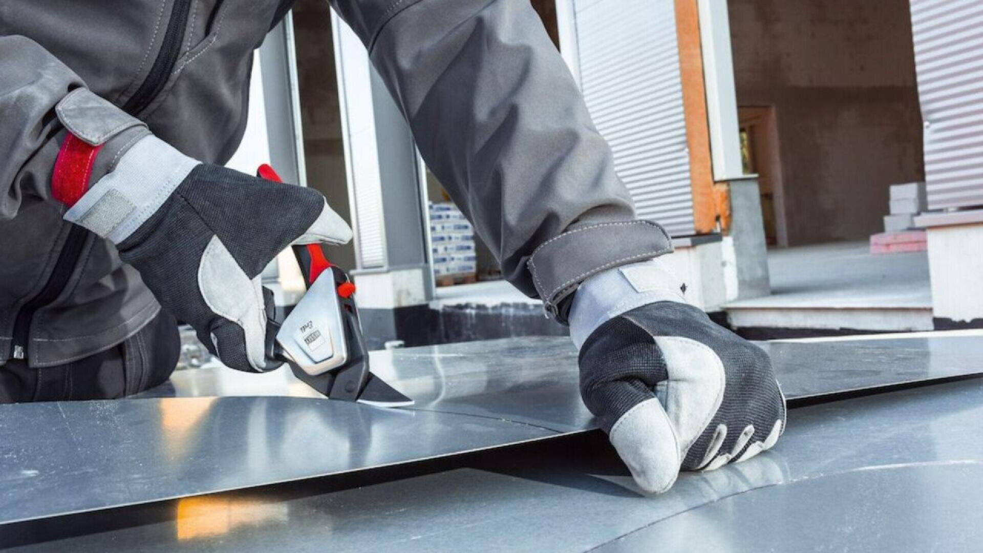 A person wearing protective gloves and a gray work jacket uses metal shears to cut a sheet of metal outdoors, with a building under construction visible in the background.