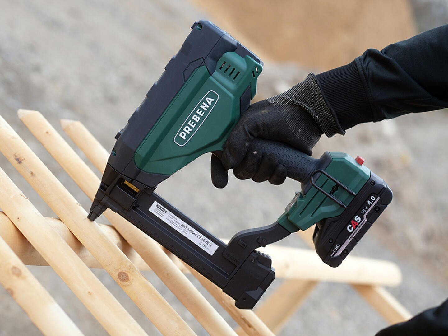Eine Person mit schwarzen Handschuhen benutzt eine grün-schwarze kabellose PREBENA-Nagelpistole, um auf einer Baustelle Holzbalken zu befestigen.