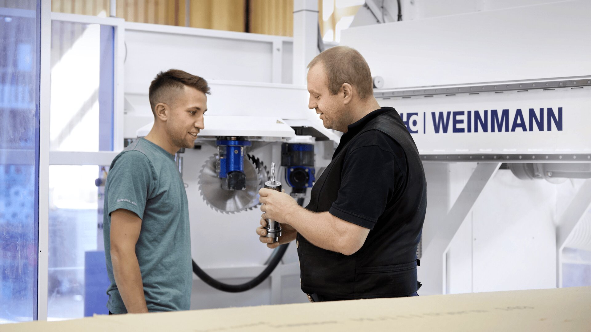 Zwei Männer stehen vor einer Industriemaschine mit der Aufschrift „WEINMANN“. Ein Mann in schwarzer Weste zeigt dem anderen Mann in blaugrünem Hemd ein kleines Metallteil. Sie scheinen sich über die Maschine oder das Gerät zu unterhalten.