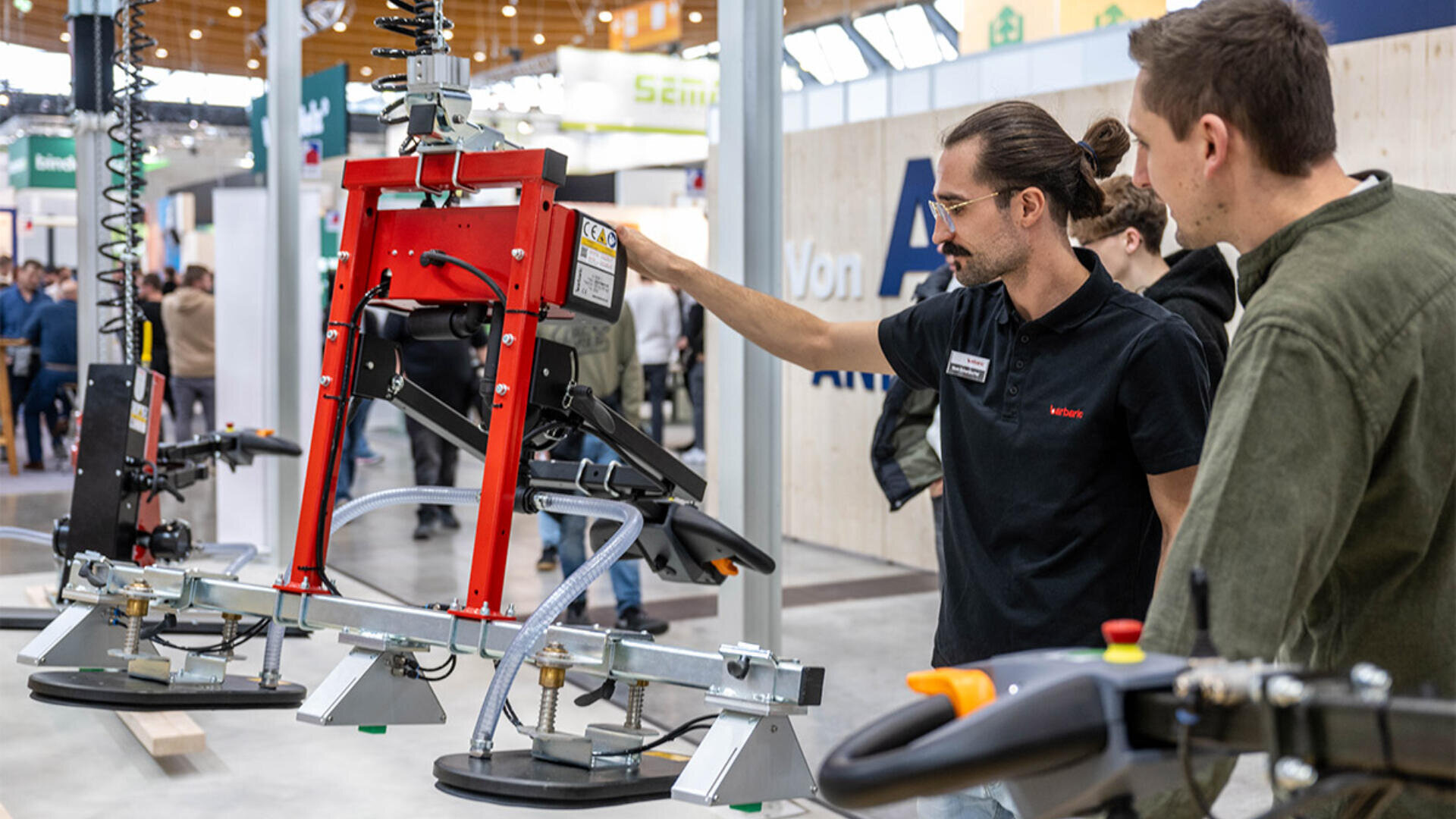Zwei Männer untersuchen auf einer Messe oder Ausstellung ein rotes Industriehebegerät mit Saugnäpfen. Ein Mann erklärt oder demonstriert dem anderen die Funktionen der Maschine.