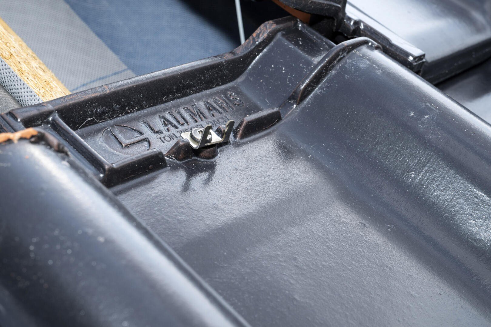Nahaufnahme einer schwarzen Metalloberfläche mit dem geprägten Wort LAUMANS und TONDACH sowie einem kleinen silbernen Metallclip in der Mitte.