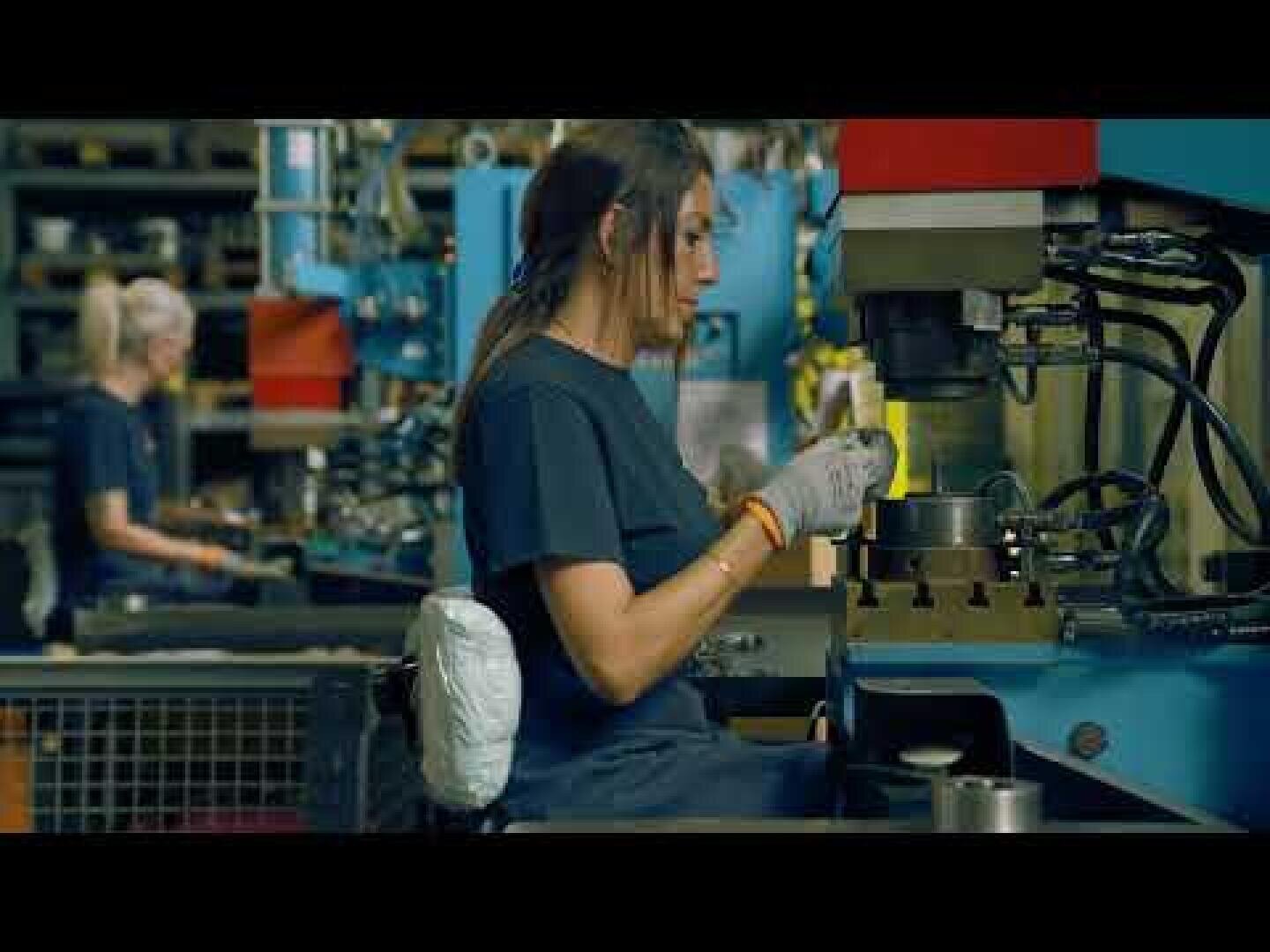 A person wearing gloves operates machinery in a factory setting, with shelves and another worker visible in the background.