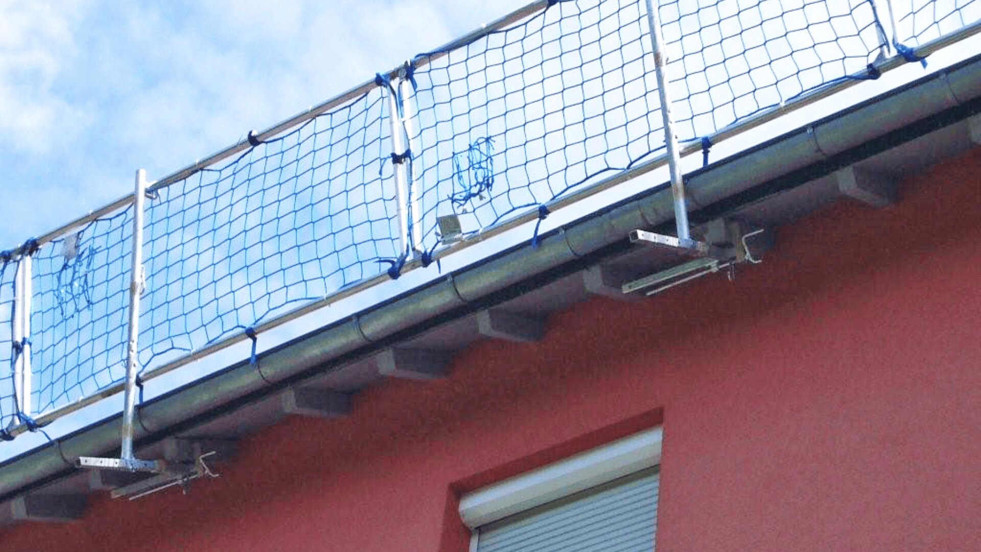 Ein Sicherheitsnetz ist entlang der Dachkante eines Gebäudes über einer roten Wand mit einem darunter liegenden geschlossenen Fenster angebracht und dient als Absturzsicherung.