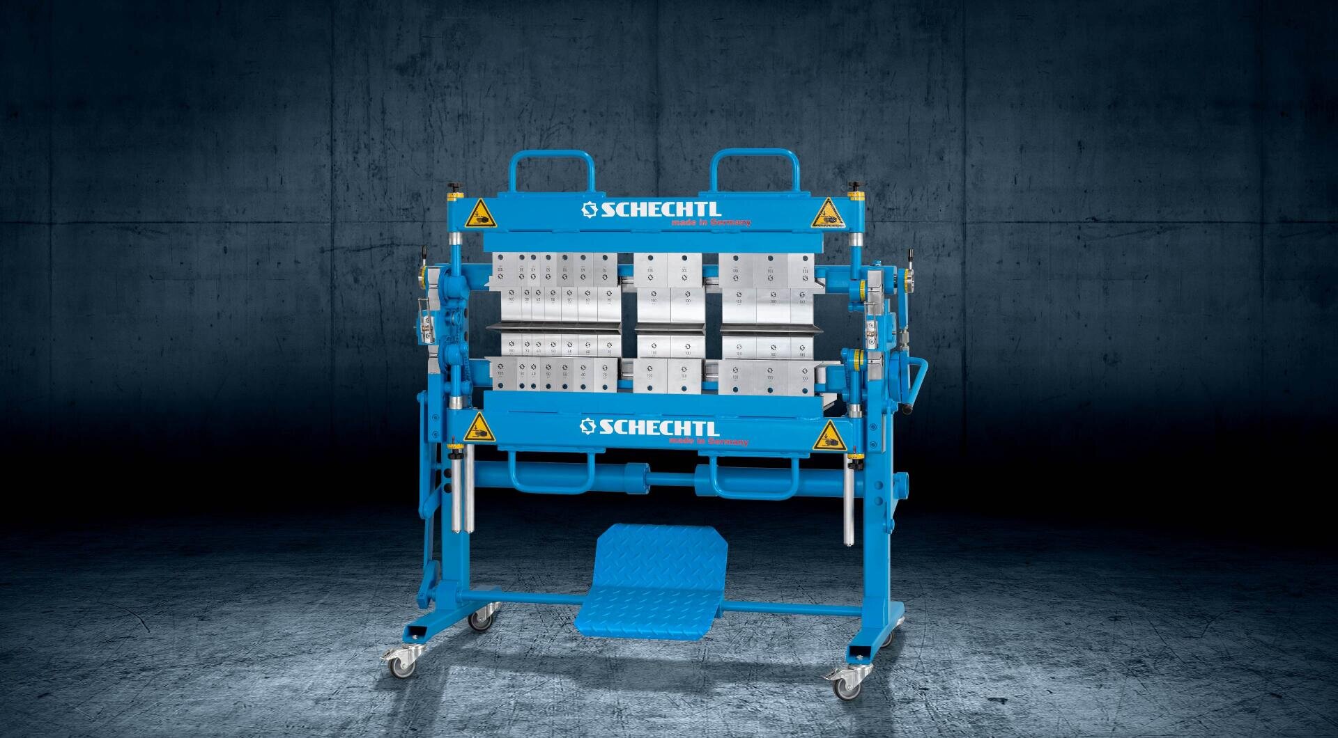 A blue Schechtl metalworking machine with visible rollers and levers stands on a wheeled frame against a dark, industrial background. Yellow caution stickers are visible on the machine.