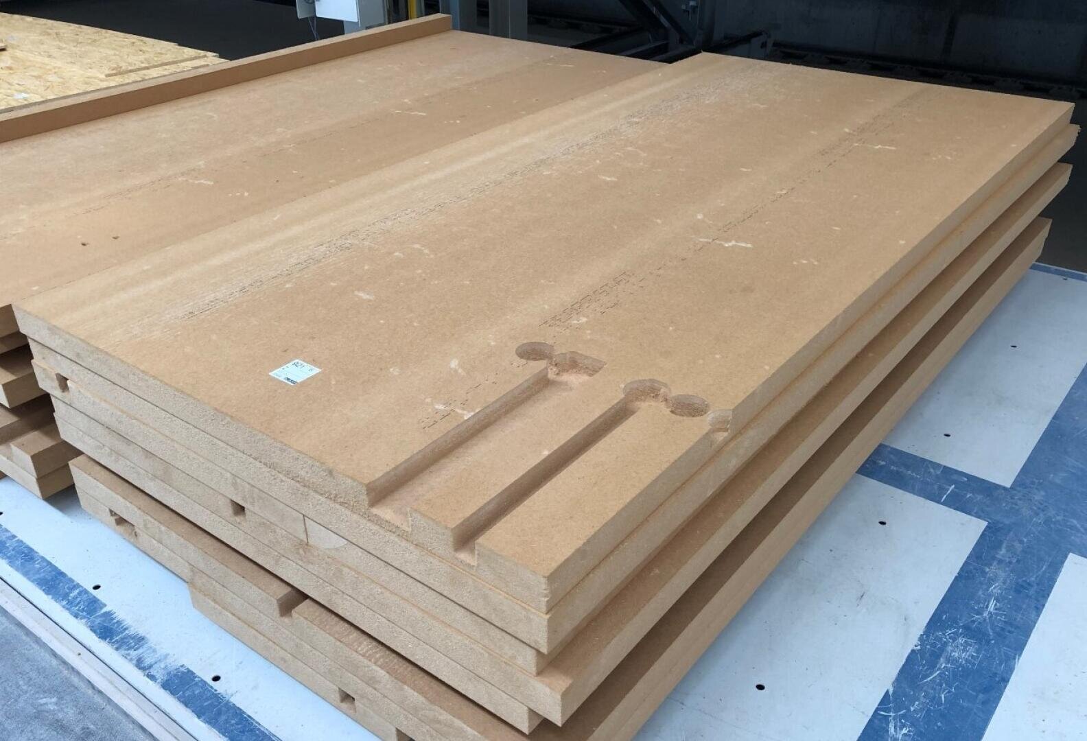 Ein Stapel großer, rechteckiger MDF-Platten mit Rillen und Kerben in den oberen Teilen, der auf einem blau-weißen Boden in einer Werkstatt oder einem Lagerhaus liegt.