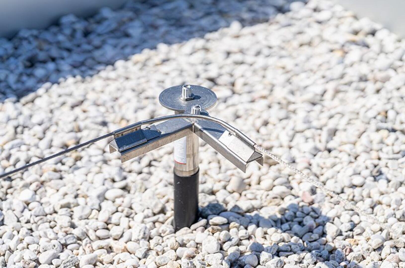 Close-up of a metal roof anchor system installed on a gravel-covered roof, with a steel cable running through the anchor for fall protection.