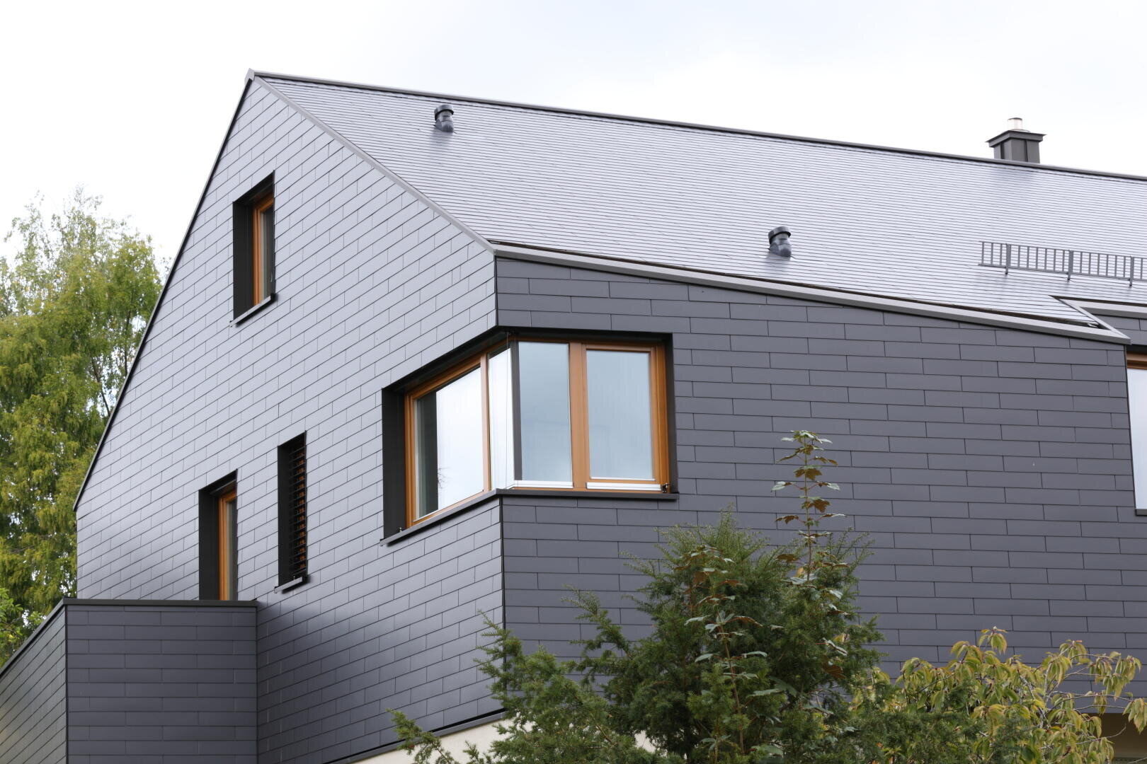 Ein modernes Haus mit dunkelgrauer Verkleidung, schrägem Dach und großen Fenstern mit Holzrahmen. Im Vordergrund sind grüne Bäume zu sehen.