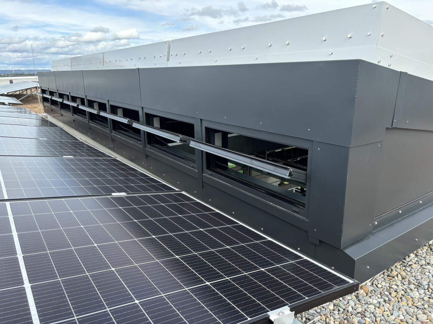 Solar panels on a roof next to a modern building with rows of small horizontal windows; the roof is covered with gravel and the sky is partly cloudy.