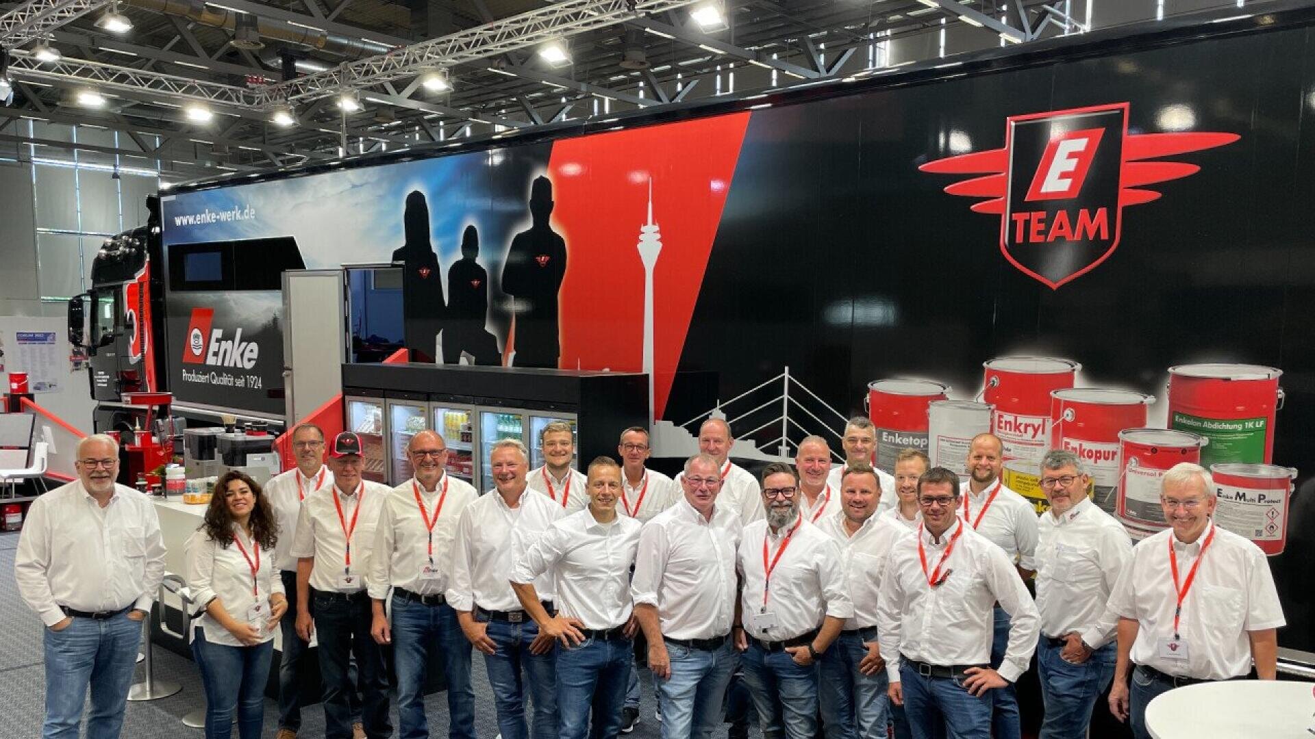 A group of people in white shirts and jeans, many wearing red lanyards, stand smiling in front of a large E Team truck with paint cans displayed and a graphic of silhouettes on its side, inside an exhibition hall.