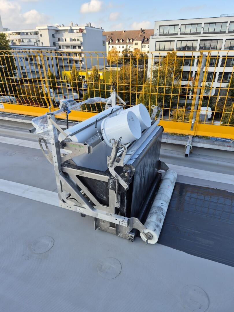Ein Bauwerkzeug mit Rollen und einem Metallrahmen hält einen weißen Farbeimer auf einem Dach. Im Hintergrund sind gelbe Sicherheitsgeländer und moderne Gebäude zu sehen.