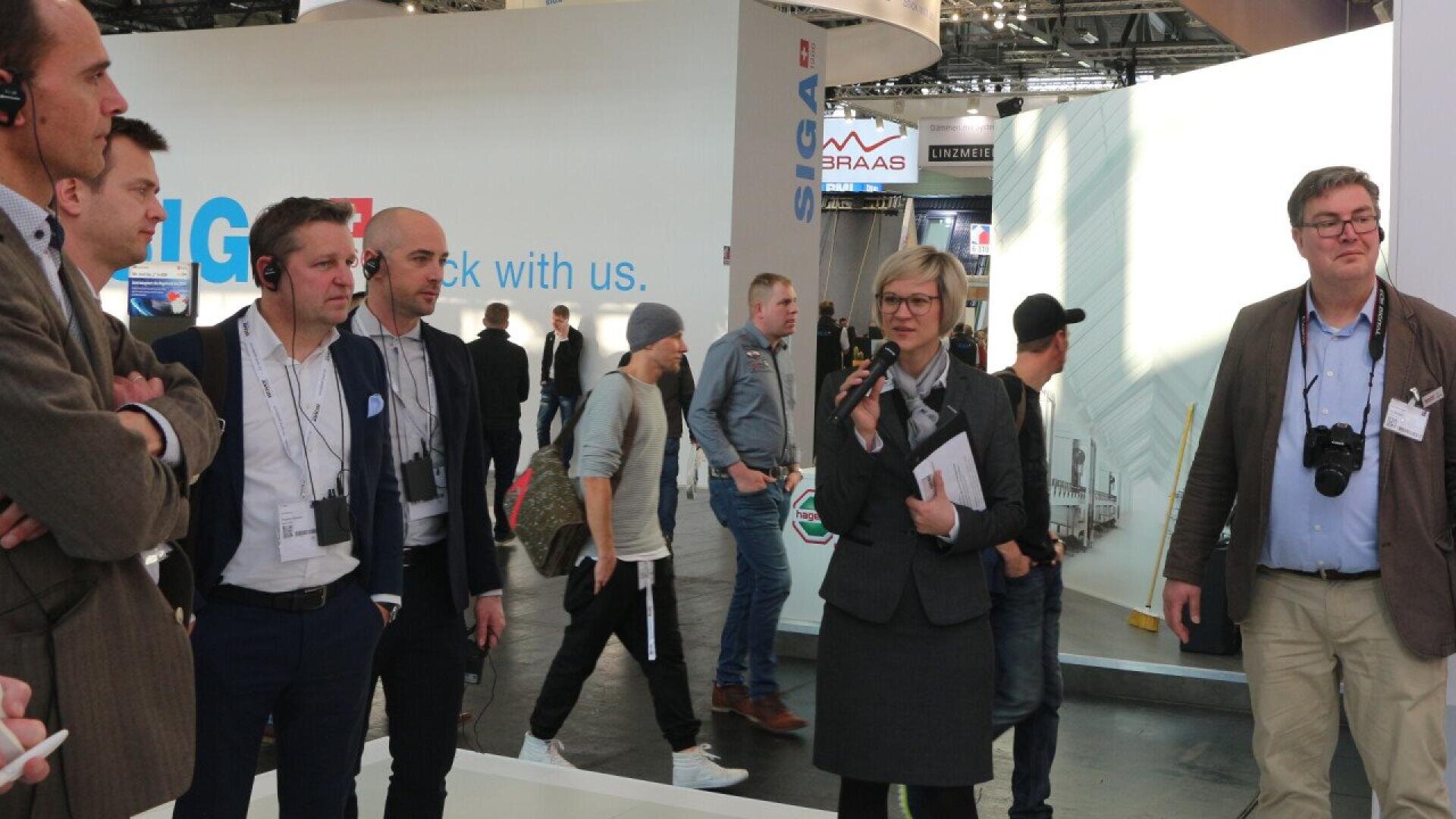 A group of people wearing business attire stand indoors at an event or expo. A woman holding a microphone speaks while others listen. Exhibition booths and banners are visible in the background.