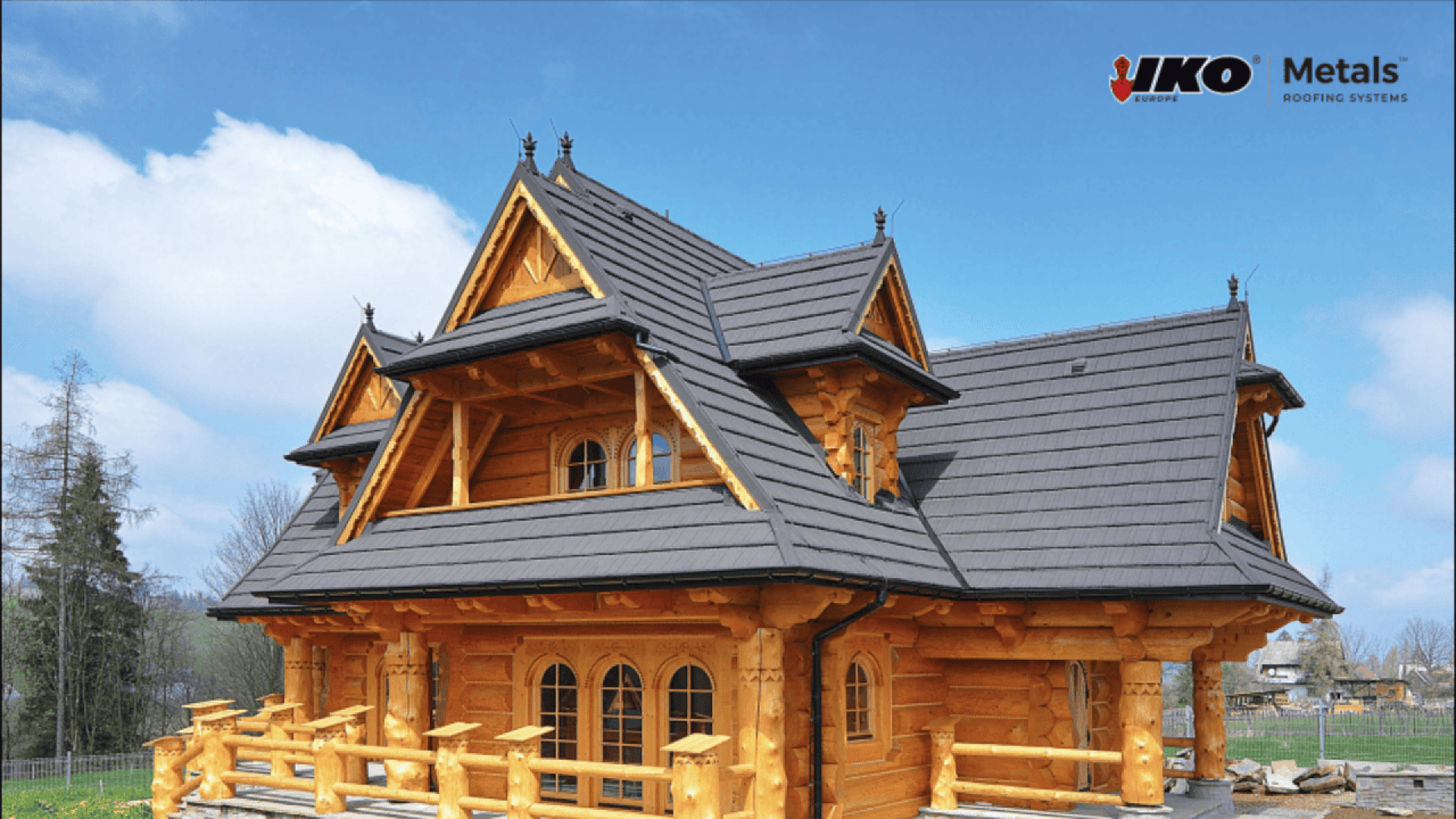 A large log cabin with intricate wooden details and steep dark gray metal roofing, set in a grassy area with trees in the background. The IKO Metals Roofing Systems logo is in the top right corner.