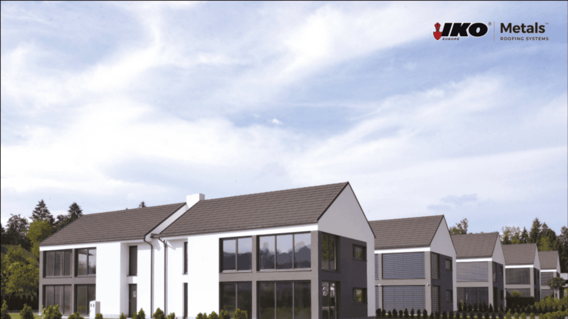 Modern residential buildings with dark gray roofs and large windows, set against a partly cloudy sky. Trees and greenery are visible in the background. The IKO Metals Roofing Systems logo appears in the top right corner.