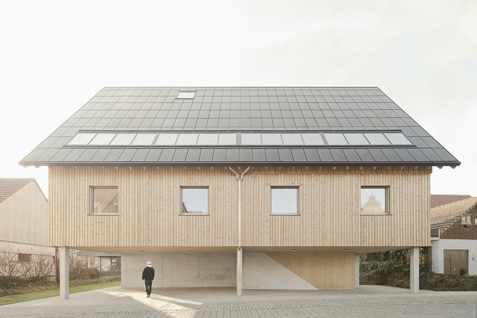 Ein modernes Holzhaus auf Stelzen mit einem großen Solarzellendach. Eine Person geht unter dem aufgeständerten Gebäude zum Eingang. Das Haus hat mehrere Fenster und einen offenen Parkplatz darunter.