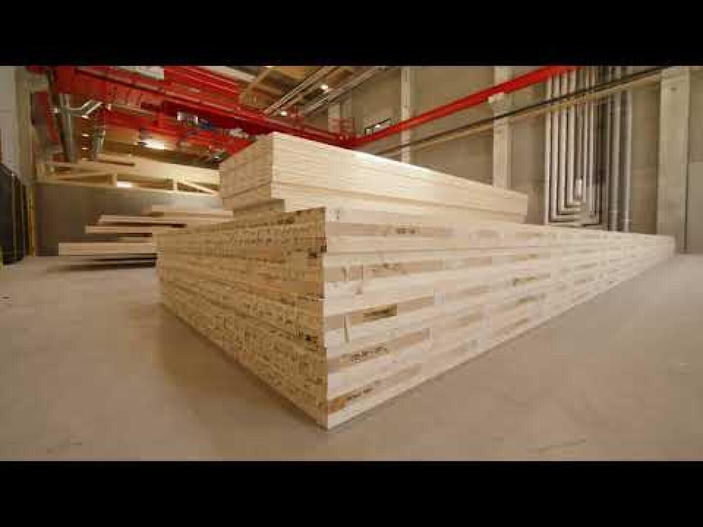 Stacks of large laminated timber beams in a spacious industrial warehouse with concrete walls, red pipes on the ceiling, and bright lighting.