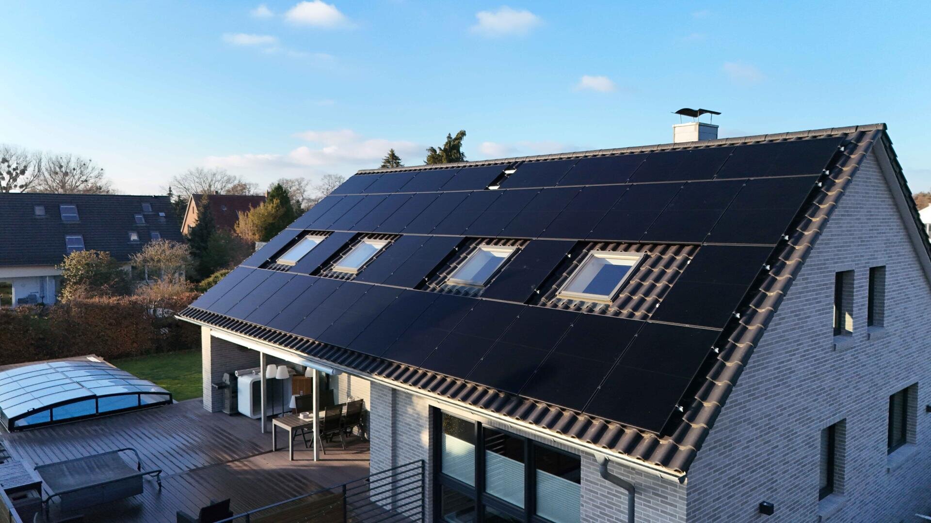 Ein modernes Haus mit einem Schrägdach, das mit schwarzen Sonnenkollektoren bedeckt ist. Auf dem Dach gibt es mehrere Oberlichter, und das Haus ist von einem Garten mit Terrassenmöbeln umgeben. Der Himmel ist meist klar mit ein paar Wolken.