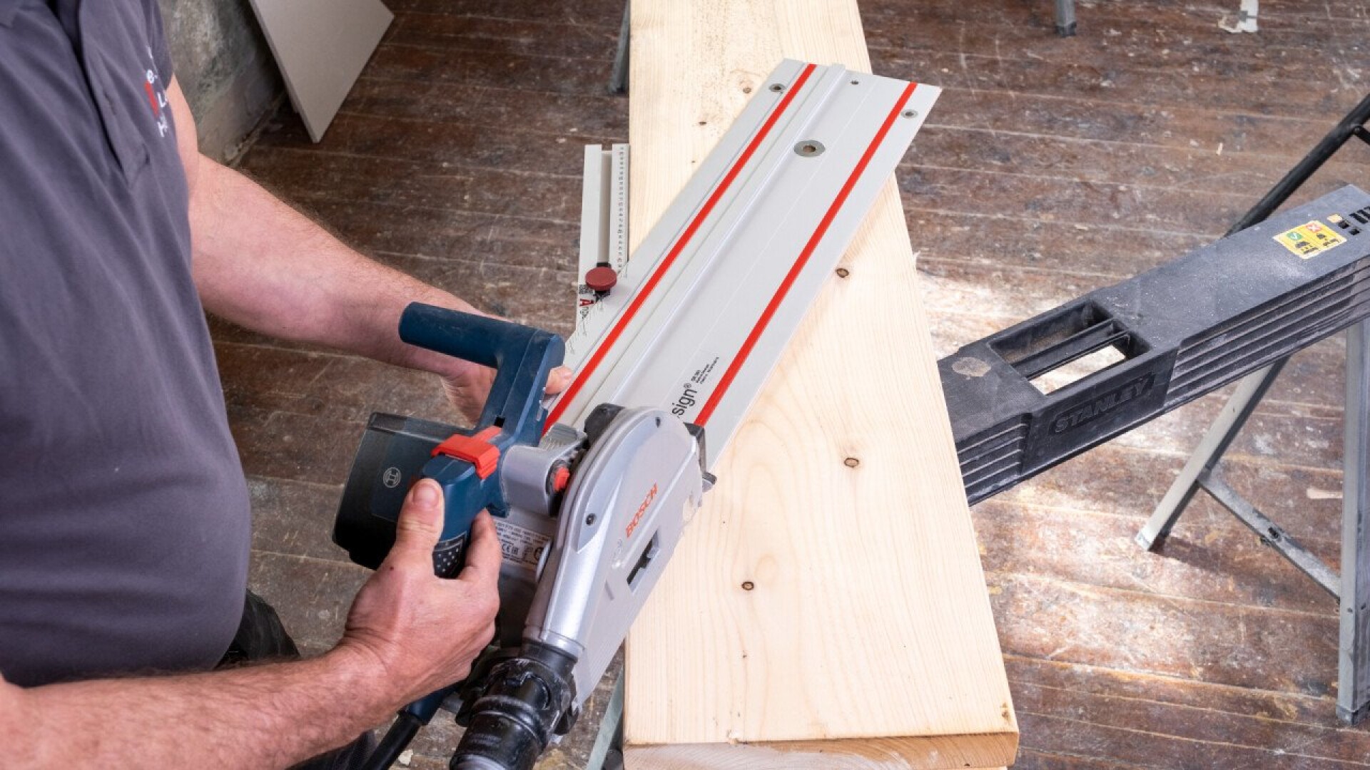 Eine Person verwendet eine Kreissäge mit Führungsschiene, um ein Stück helles Holz zu schneiden, das von einem Sägebock auf einem Holzboden gestützt wird.