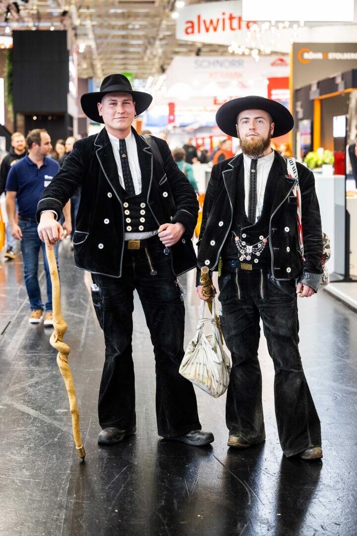 Zwei Männer in traditioneller schwarzer Zimmermannskleidung, darunter breitkrempige Hüte, Westen und Schlaghosen, stehen in einem Haus. Einer hält einen Holzstab, der andere trägt eine weiße Stofftasche. Im Hintergrund sind Menschen und Stände zu sehen.