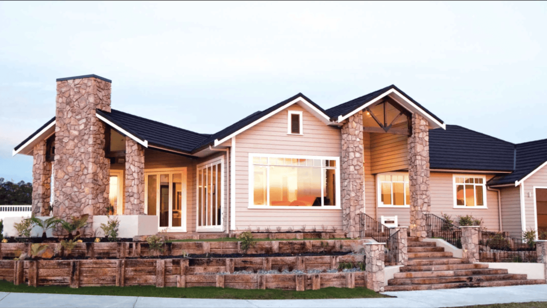Modern, single-story house with stone accent walls, large windows, and a dark roof. Raised garden beds and stairs lead to the front entrance, with outdoor lights illuminating the home at dusk.