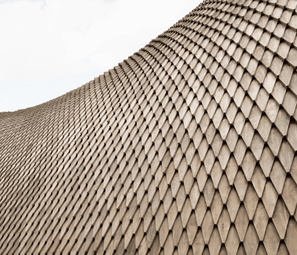 Curved architectural structure covered with overlapping, diamond-shaped wooden shingles, creating a textured, scale-like pattern on the surface.