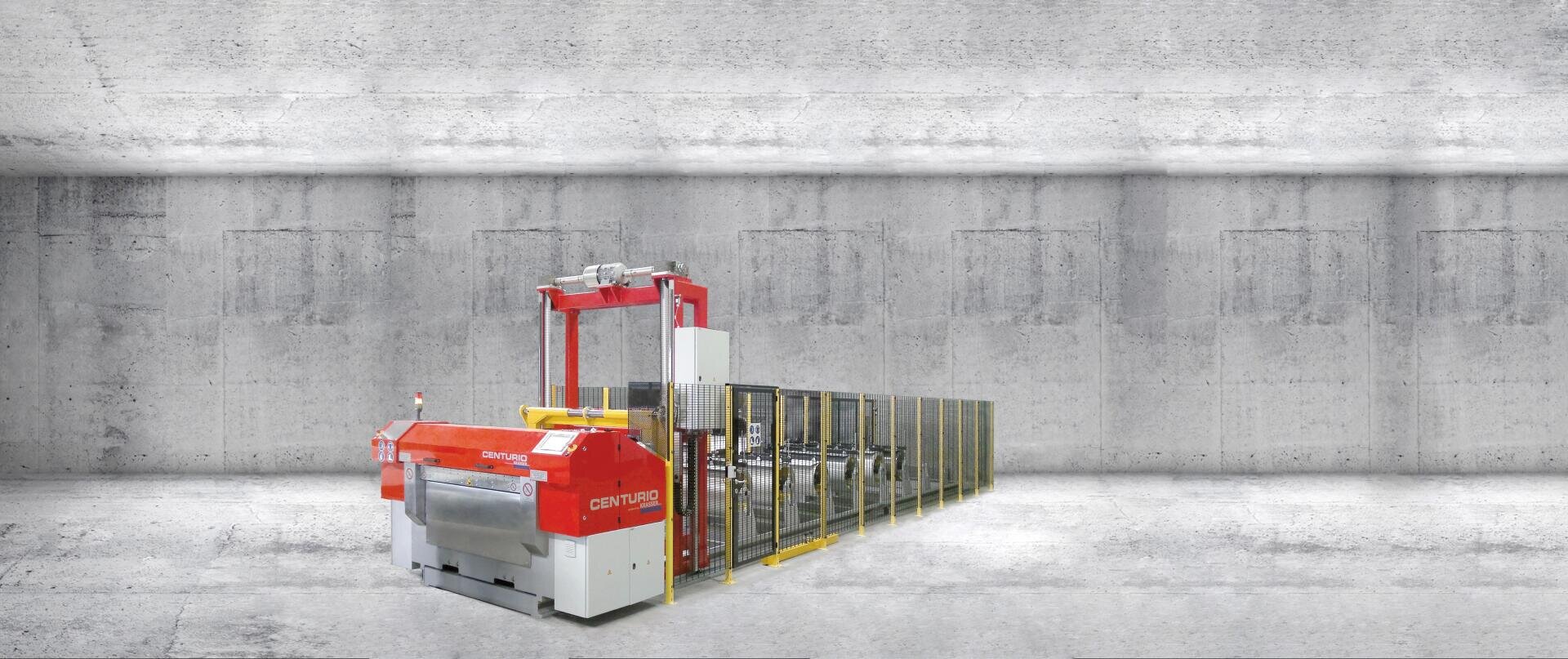 Eine große Industriemaschine mit rot-silberner Außenfassade steht in einem geräumigen, minimalistischen Betonraum. Die Maschine ist mit einem Fördersystem und Schutzbarrieren entlang der gesamten Länge ausgestattet.