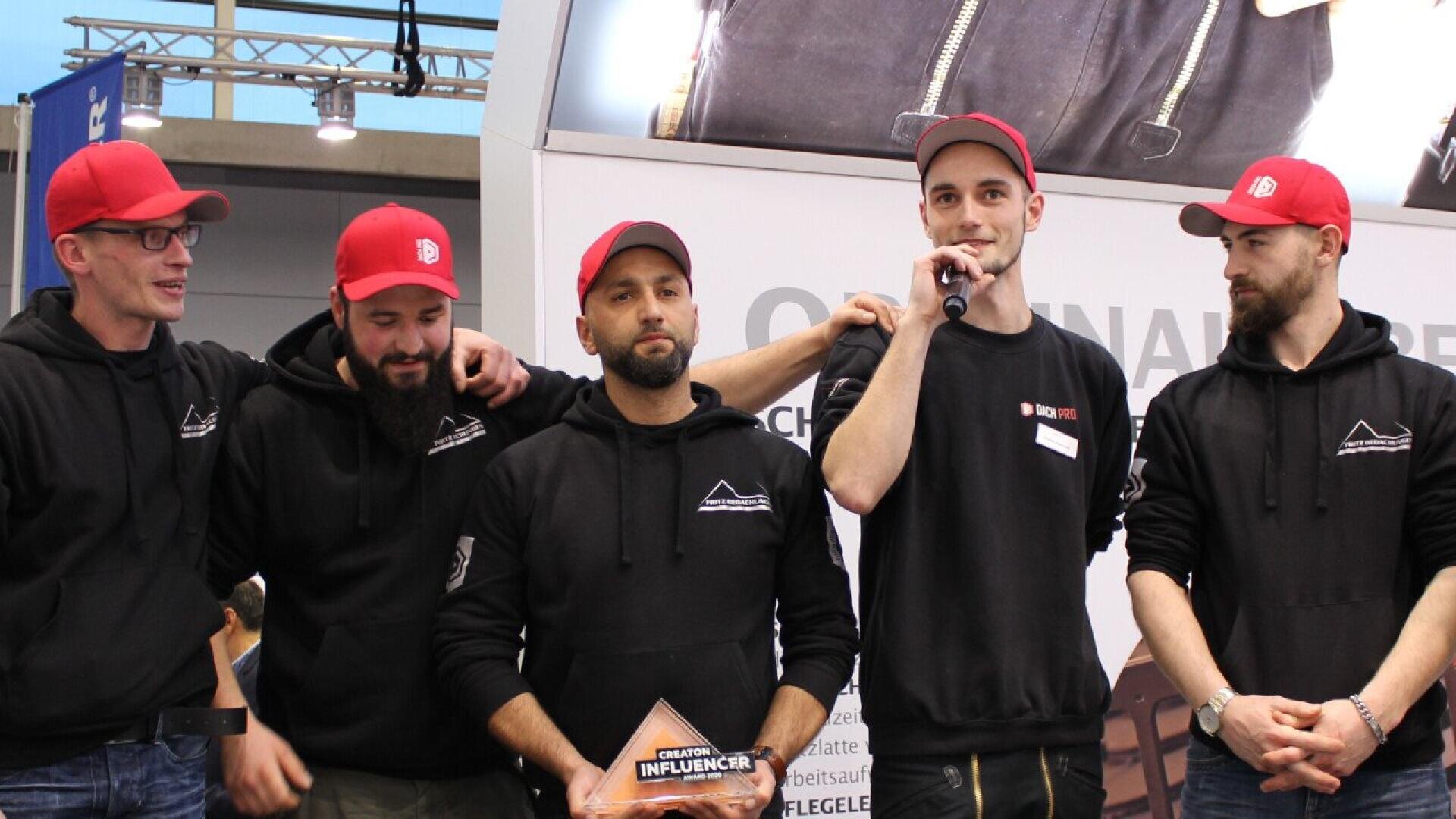 Five men in matching black hoodies and red caps stand together on a stage; one is holding a microphone and speaking, another holds a glass award. They appear to be at an indoor event or competition.