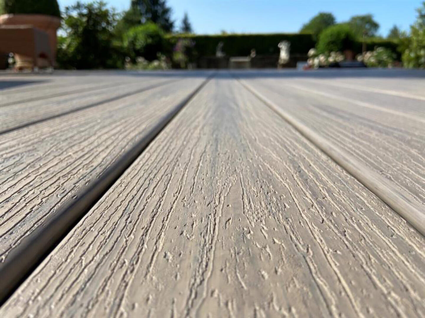 Nahaufnahme, Untersicht von strukturierten Holzterrassenbrettern im Freien, mit einem unscharfen Hintergrund aus Grün, Pflanzen und einem klaren blauen Himmel an einem sonnigen Tag.