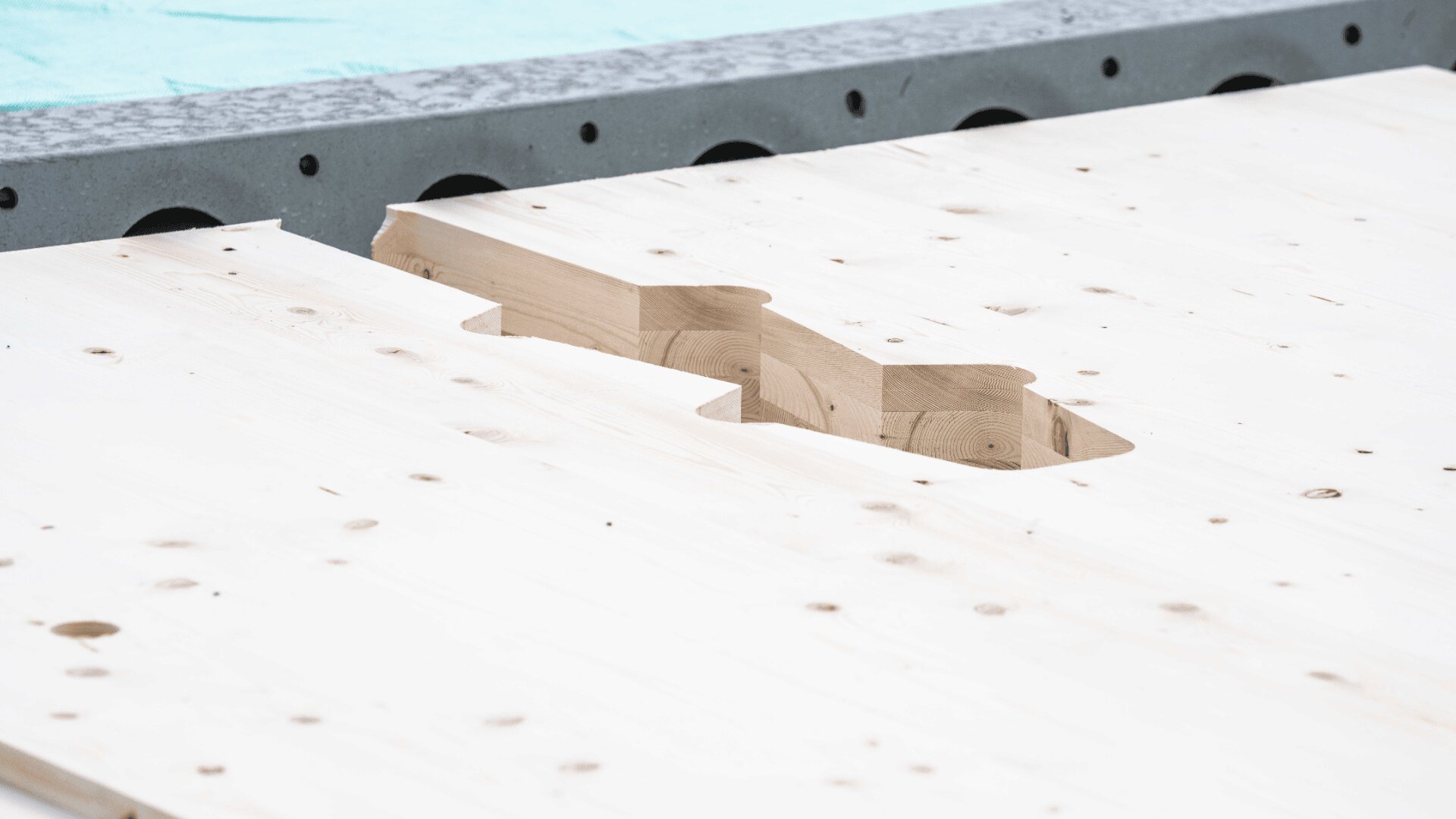 Eine Nahaufnahme von unbearbeiteten Holzplatten mit einer präzise geschnittenen, ineinandergreifenden Verbindung, die auf einem Betonsockel auf einer Baustelle ruhen.