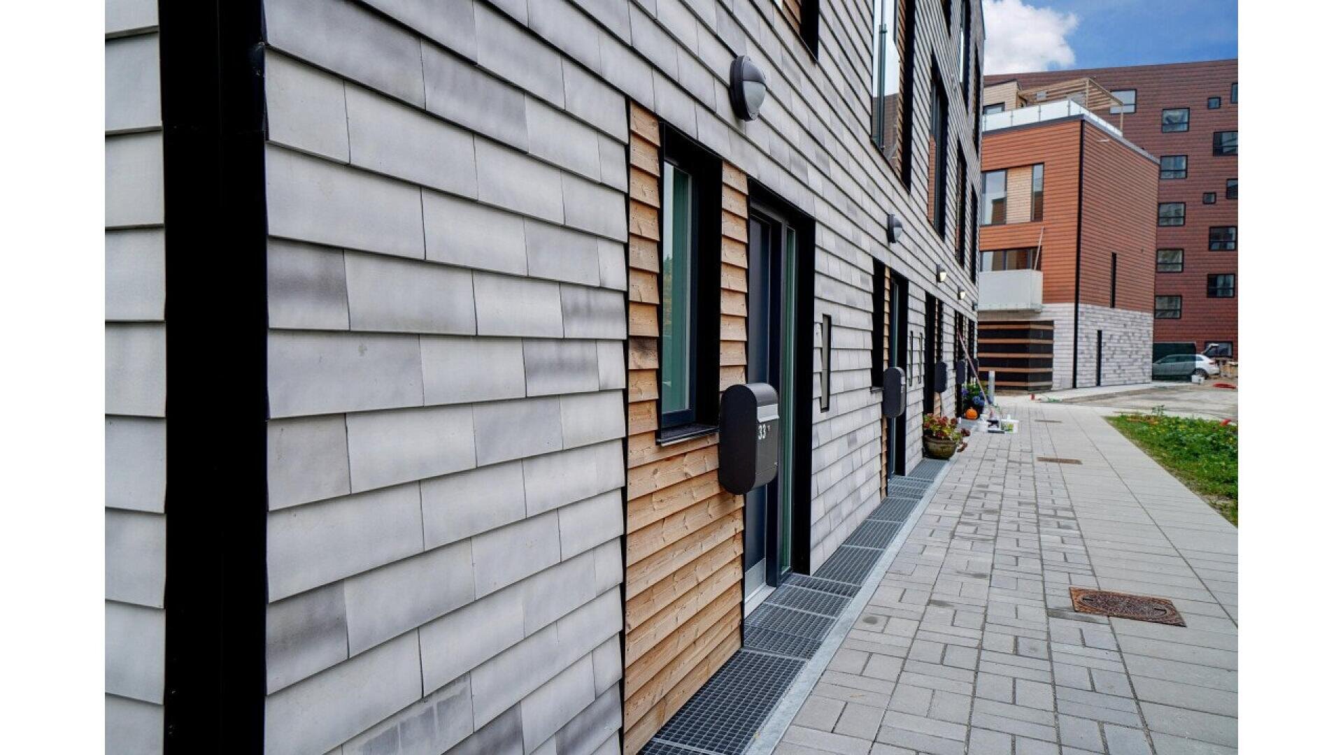 A modern apartment building with gray and natural wood siding, black doors with electronic locks, and a paved walkway along the front. Other apartment buildings and green grass are visible in the background.