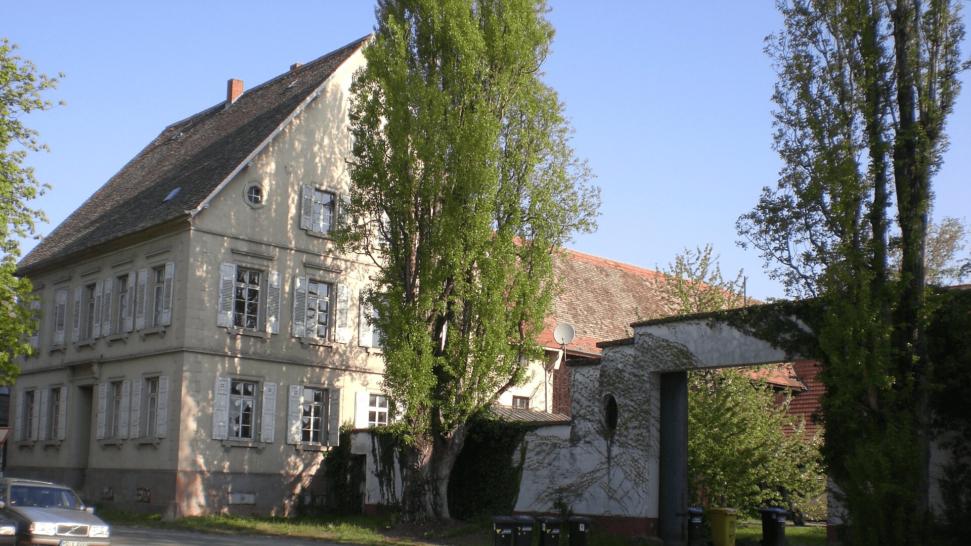 Ein großes, altmodisches beiges Haus mit schrägem Dach wird teilweise von hohen Bäumen beschattet. In der Nähe steht ein weißer Torbogen, und das Sonnenlicht beleuchtet Teile des Gebäudes und die grüne Umgebung.