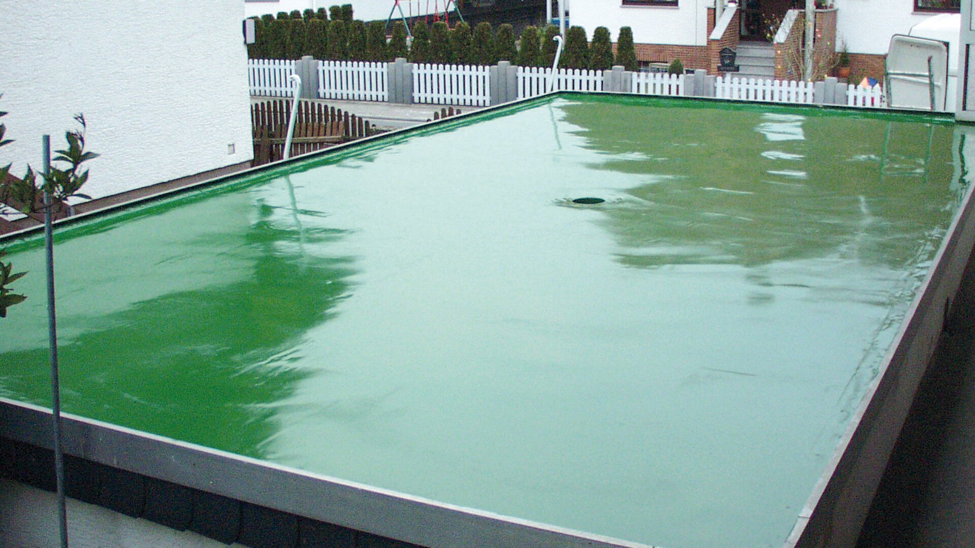 Ein Flachdach mit einer glatten, glänzenden, grünen, wasserdichten Beschichtung, in der sich die umliegenden Häuser und weißen Zäune in einem Wohngebiet widerspiegeln.