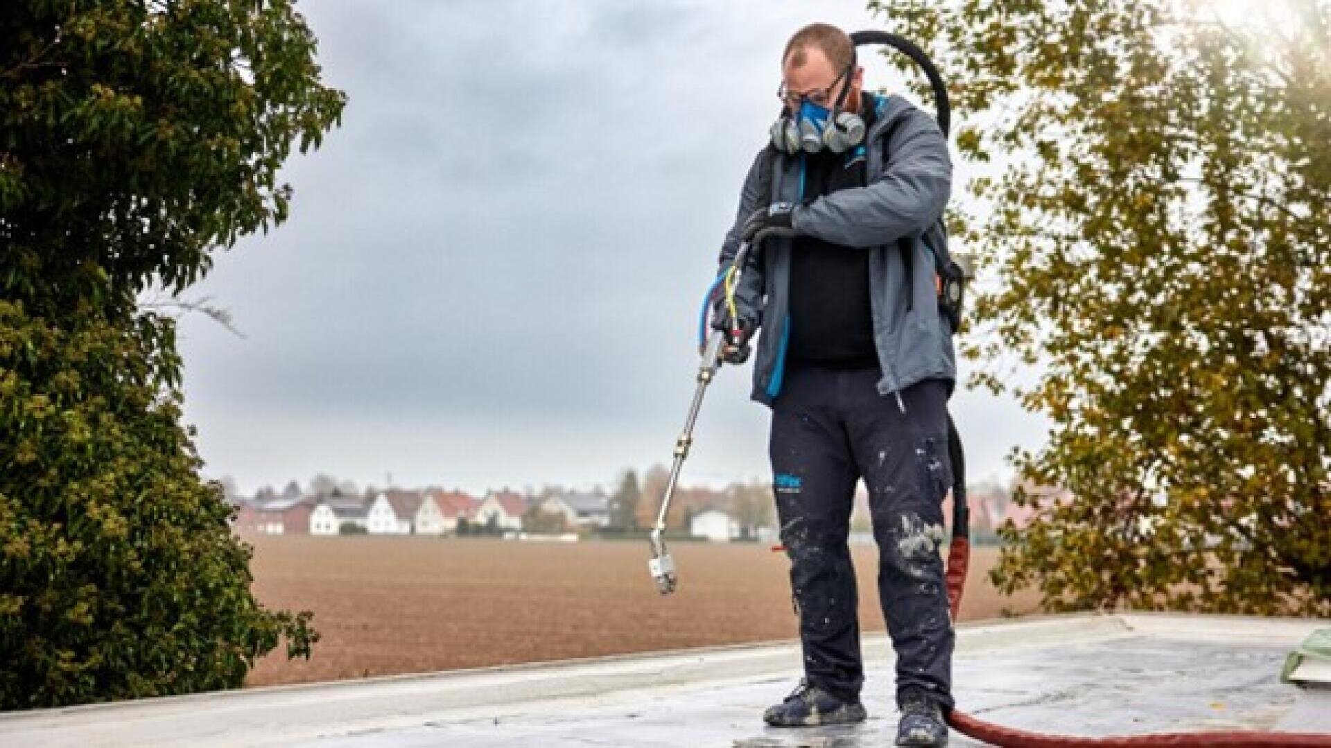 Eine Person mit Atemschutzmaske und Schutzkleidung steht im Freien und benutzt ein Sprühgerät auf einer ebenen Fläche, im Hintergrund sind Bäume und ein Feld unter einem bewölkten Himmel zu sehen.