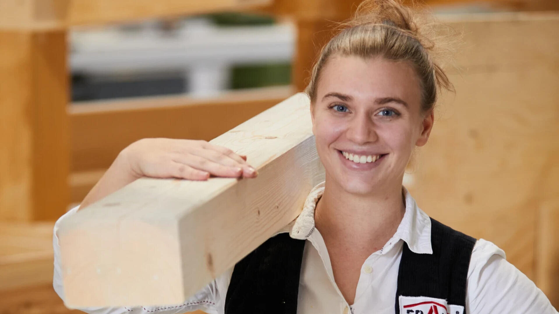 Eine lächelnde Frau in Weste und weißem Hemd hält in einer Werkstatt einen großen Holzbalken auf ihrer Schulter.