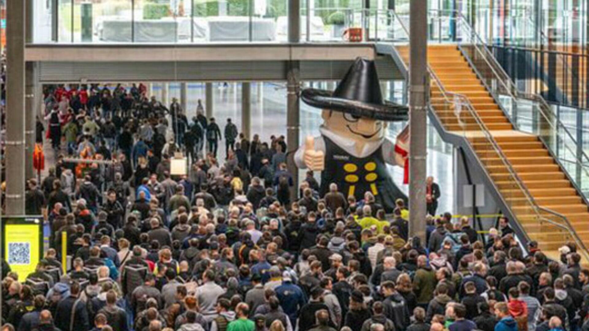 A large crowd gathers indoors near a giant inflatable figure wearing a black hat and glasses, next to a staircase and large windows in a spacious corridor.