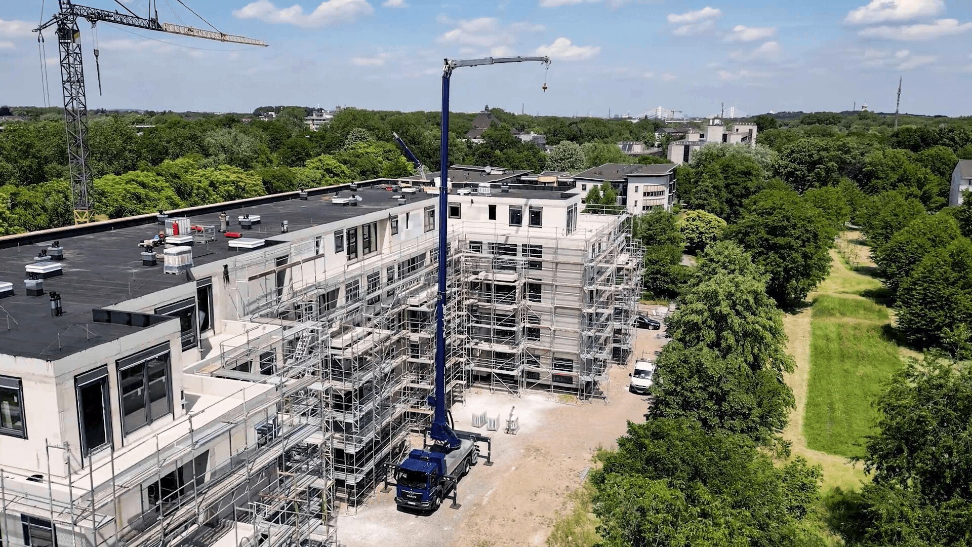 Eine Baustelle mit einem großen Gebäude, das renoviert wird und von Gerüsten umgeben ist. Ein blauer Kranwagen steht neben dem Gebäude, und auf der rechten Seite befinden sich Bäume und eine grüne Wiese unter blauem Himmel.