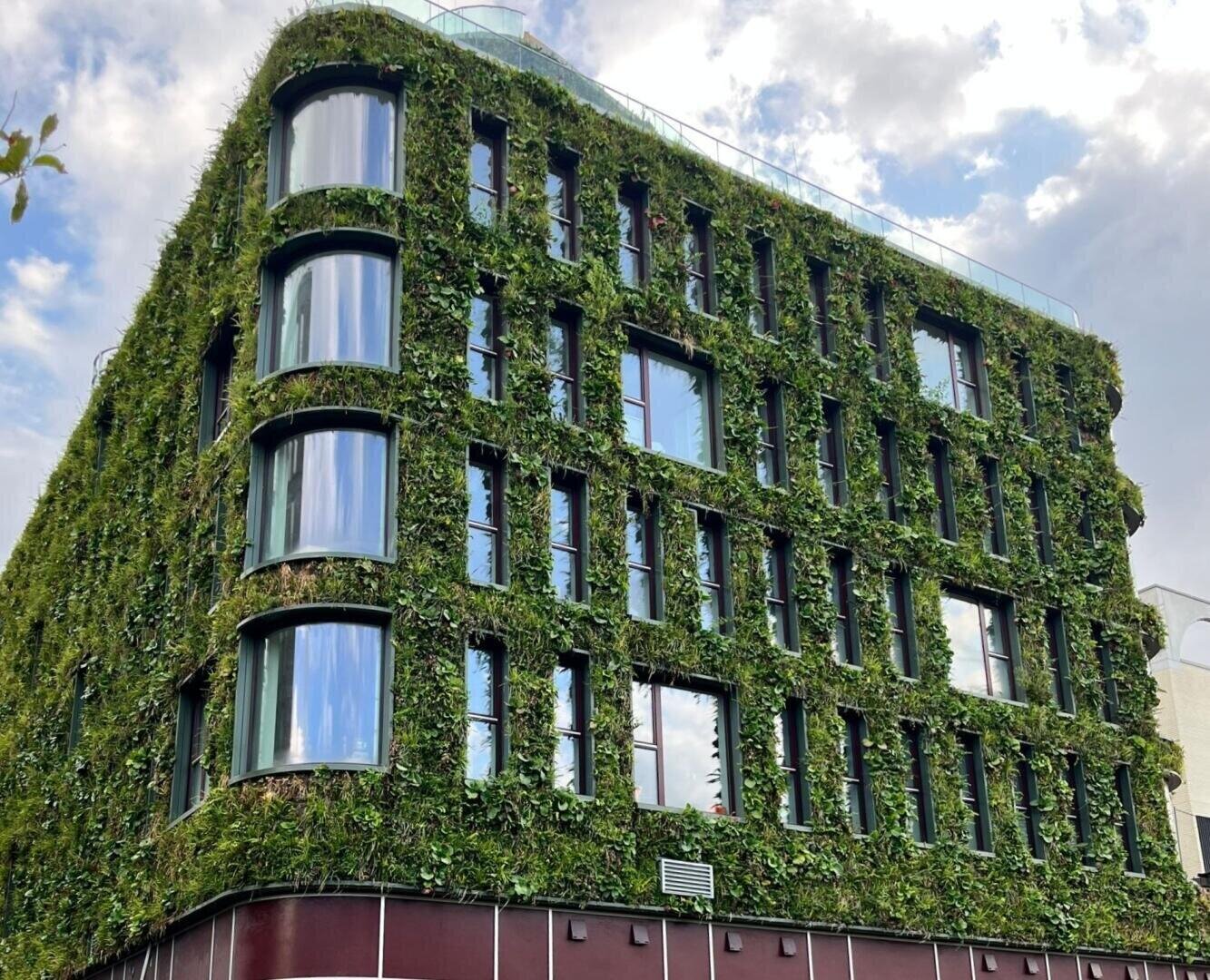 A modern building covered with green plants on its exterior walls, featuring large windows and a rounded glass corner, set against a partly cloudy sky.