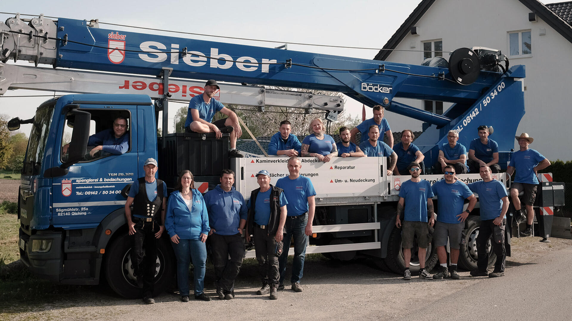 A group of workers in blue shirts pose together in front of a large Sieber-branded crane truck parked on a residential street, with a white house and greenery in the background.